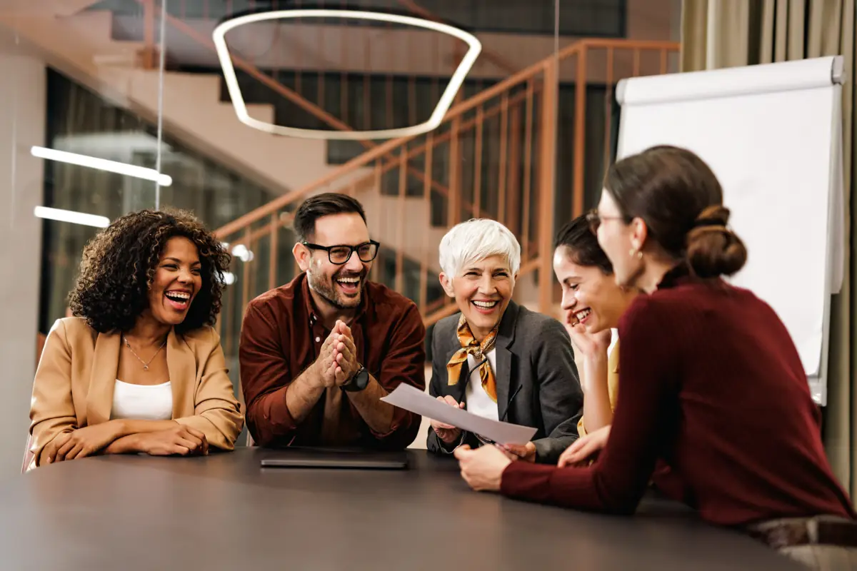 Five smiling colleagues of different backgrounds laughing together around a meeting table